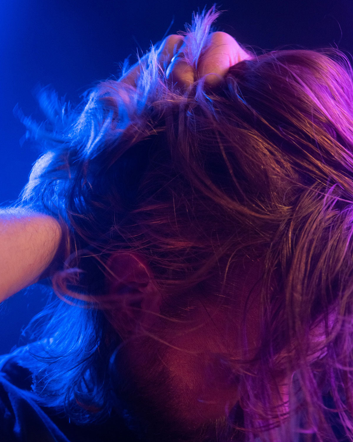 Eric running hands through his long hair highlighted under vibrant lighting. His head is angled down towards the right.