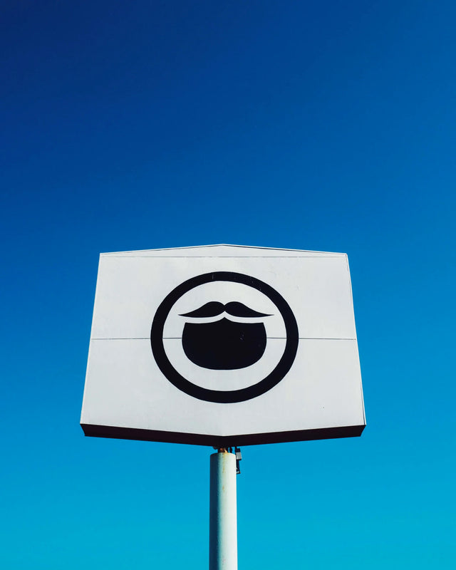 Beardbrand Barbershop signage is a white hexagon shape stretched to nearly be a square and has the Beardbrand circle beard icon on the front in black. The sign is on top of a metal post and is in front of a cloudless electric blue sky in Austin, Texas. The sky is a bright gradient of light blues from the bottom to darker hues of blue near the top left corner of the image. 