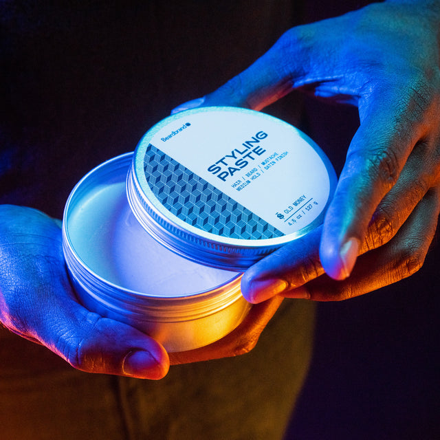 A slightly open canister of Beardbrand Styling Paste held in a person's hands with vibrant lighting so the top lid label is showing.
